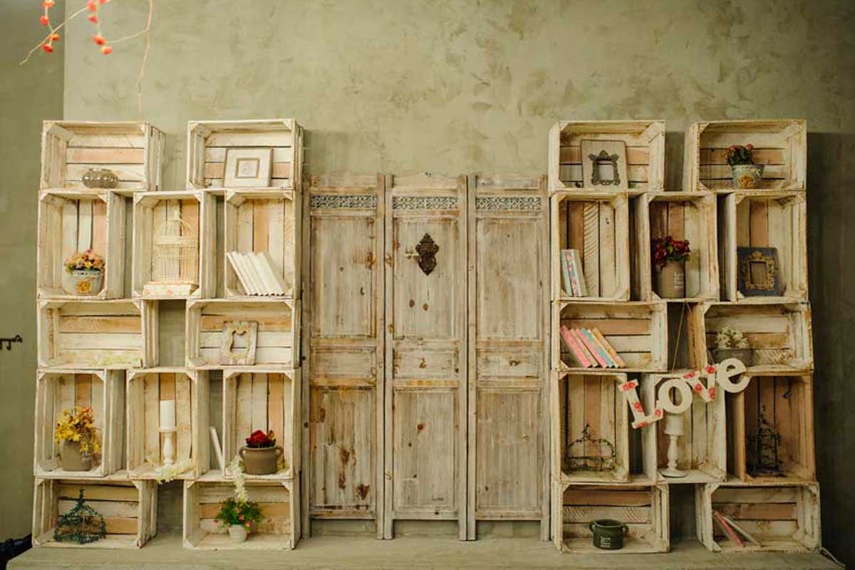 Libreria realizzata con cassette di legno dipinte di bianco, decorata con libri, cornici e piccoli oggetti, su sfondo di parete shabby e pannelli vintage. Stile rustico e creativo.