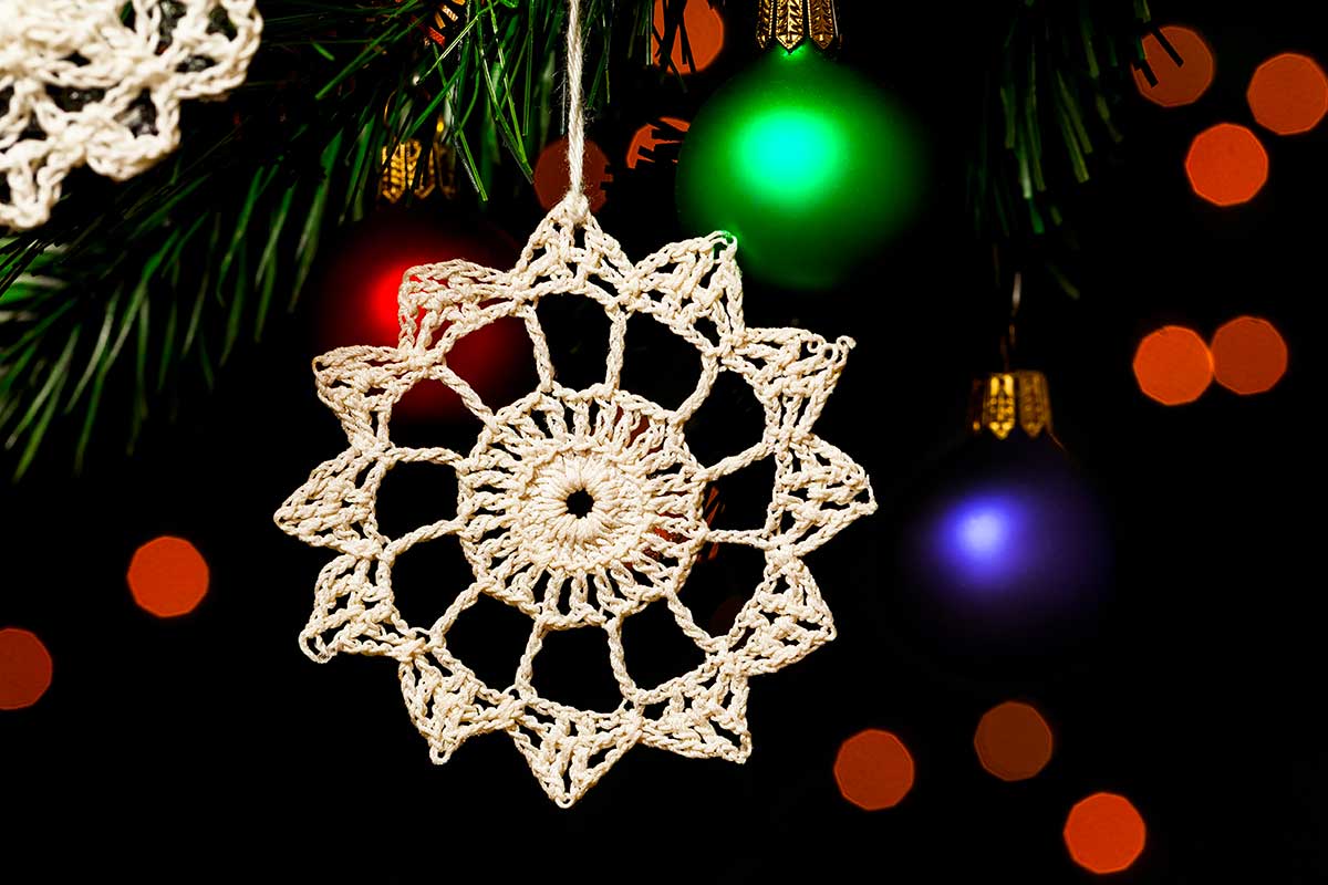 Centrino all’uncinetto appeso a un ramo di albero di Natale, illuminato da luci soffuse e palline colorate sullo sfondo. Decorazione handmade dal sapore vintage.