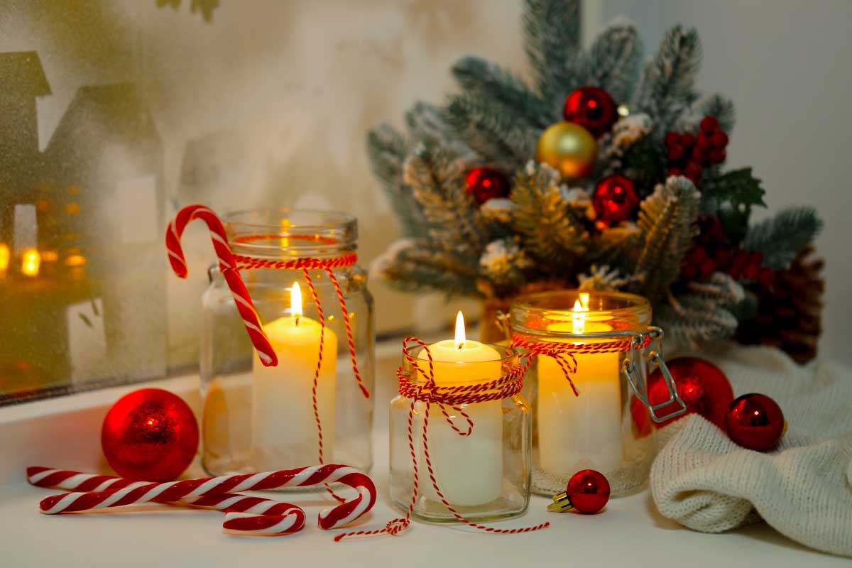 Barattoli di vetro decorati con spago rosso e bianco usati come portacandele, circondati da bastoncini di zucchero, palline natalizie e rami innevati su un davanzale invernale.
