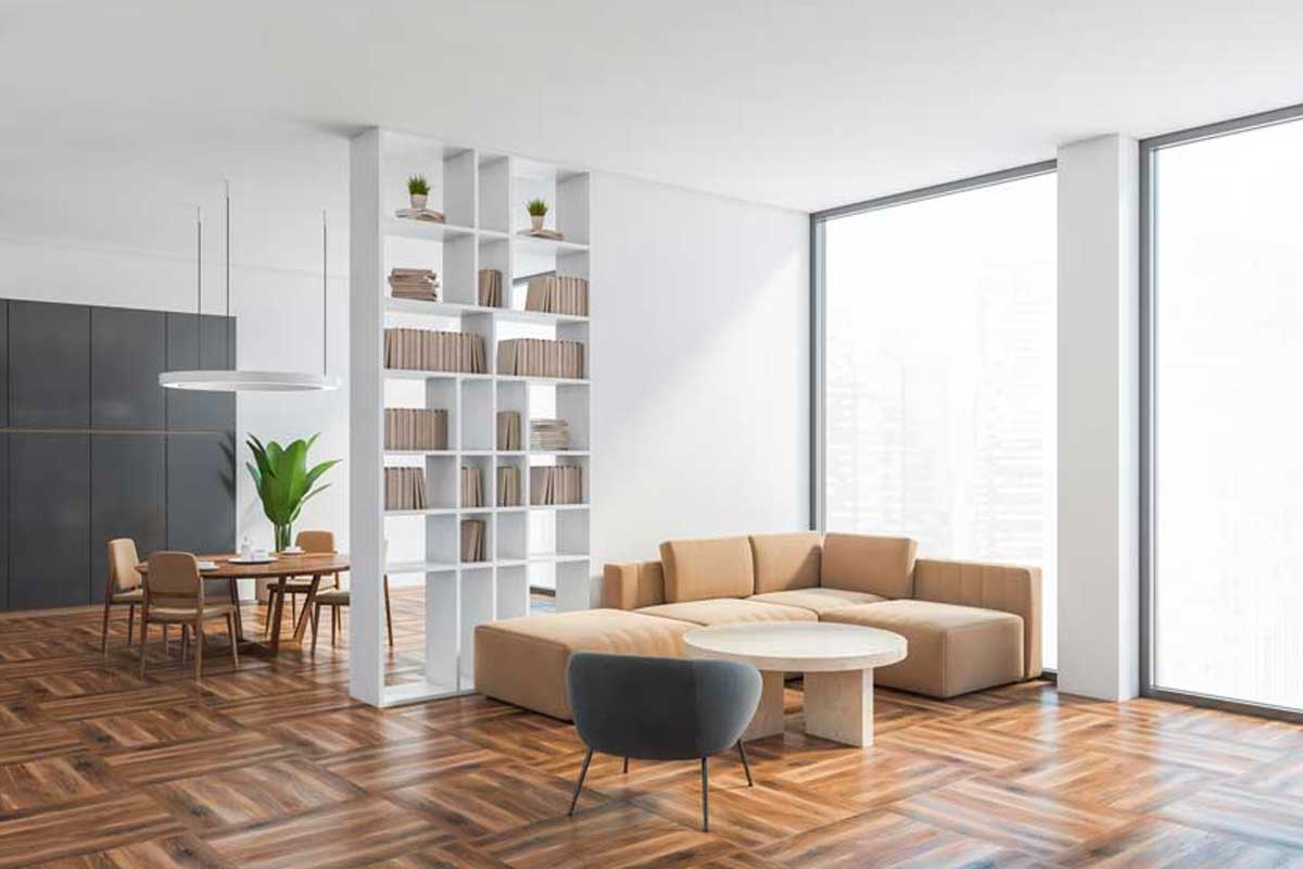 Open space luminoso con grande libreria bianca che separa zona cucina e soggiorno. Parquet caldo, tavolo da pranzo in legno chiaro, divano beige e ampie vetrate che portano luce naturale. Atmosfera ordinata e accogliente.