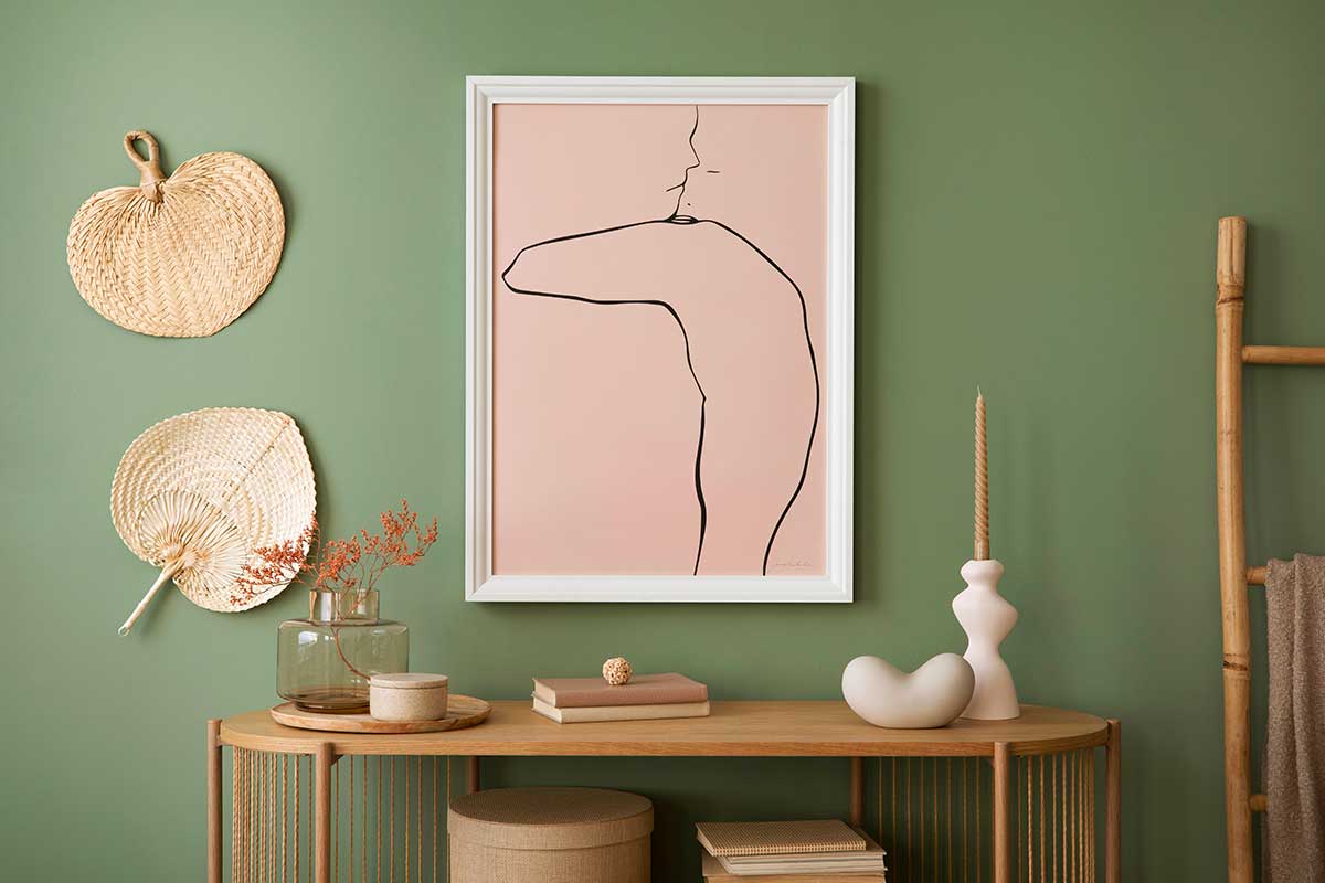 Angolo di soggiorno moderno con parete verde salvia, consolle in legno chiaro decorata con libri, vasi e candela. Sopra, quadro minimal su fondo rosa e ventagli in paglia appesi alla parete.