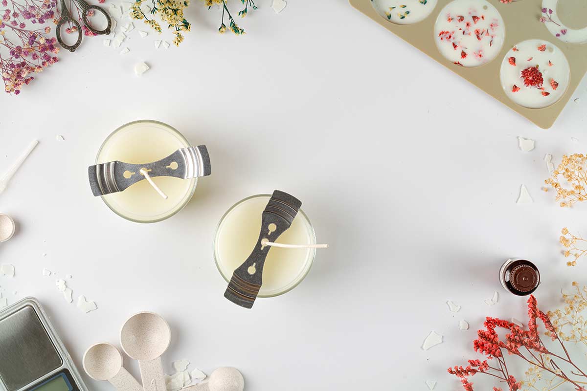 Barattoli di vetro trasformati in candele fai da te, stoppini fissati con supporti, intorno strumenti da cucina e decorazioni floreali essiccate su fondo bianco.