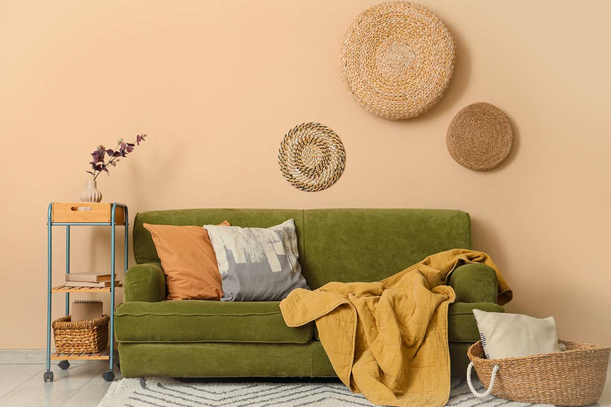Divano verde oliva in soggiorno con cuscini e coperta senape, cestino in vimini con manici sul tappeto, decorazioni a parete con ceste intrecciate tonde e mobiletto laterale con vaso e libri.