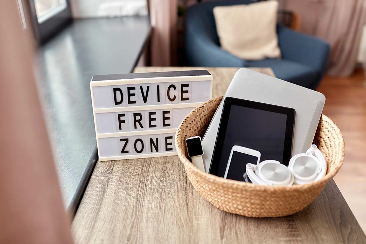 Cestino in vimini naturale su tavolo in soggiorno, usato come contenitore per dispositivi elettronici (tablet, smartphone, cuffie, laptop) accanto a un’insegna con la scritta “Device free zone”.