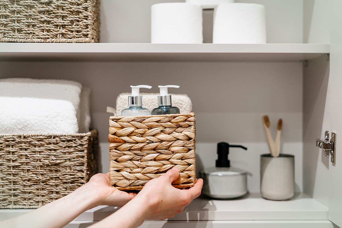 Mani che sistemano un cestino intrecciato su uno scaffale in bagno, con dispenser di sapone, asciugamani bianchi, rotoli di carta e accessori ordinati.