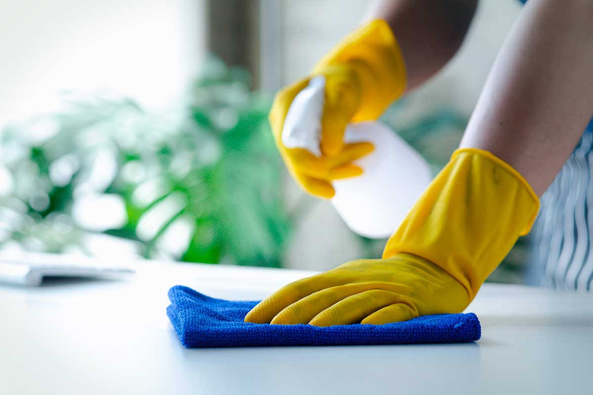 detergenti probiotici: mani con guanti gialli che puliscono una superficie bianca con un panno in microfibra blu e spray, sullo sfondo piante verdi sfocate, foto © kamiphotos - stock adobe