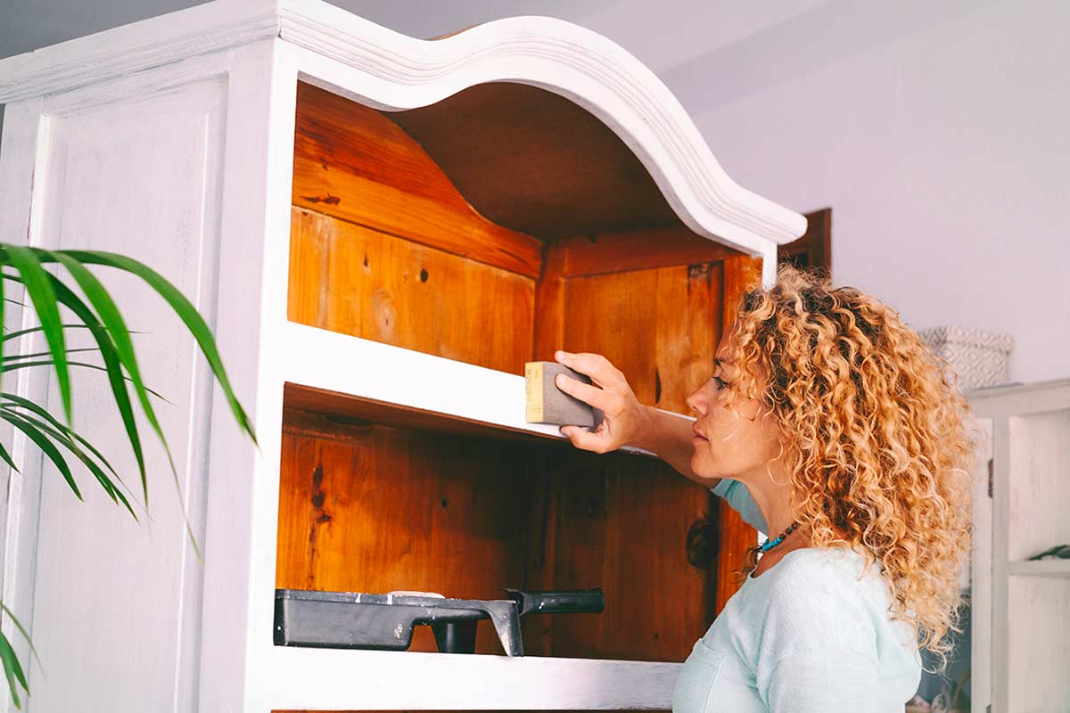 Donna che carteggia una credenza in legno chiaro, preparandola per una trasformazione fai da te.