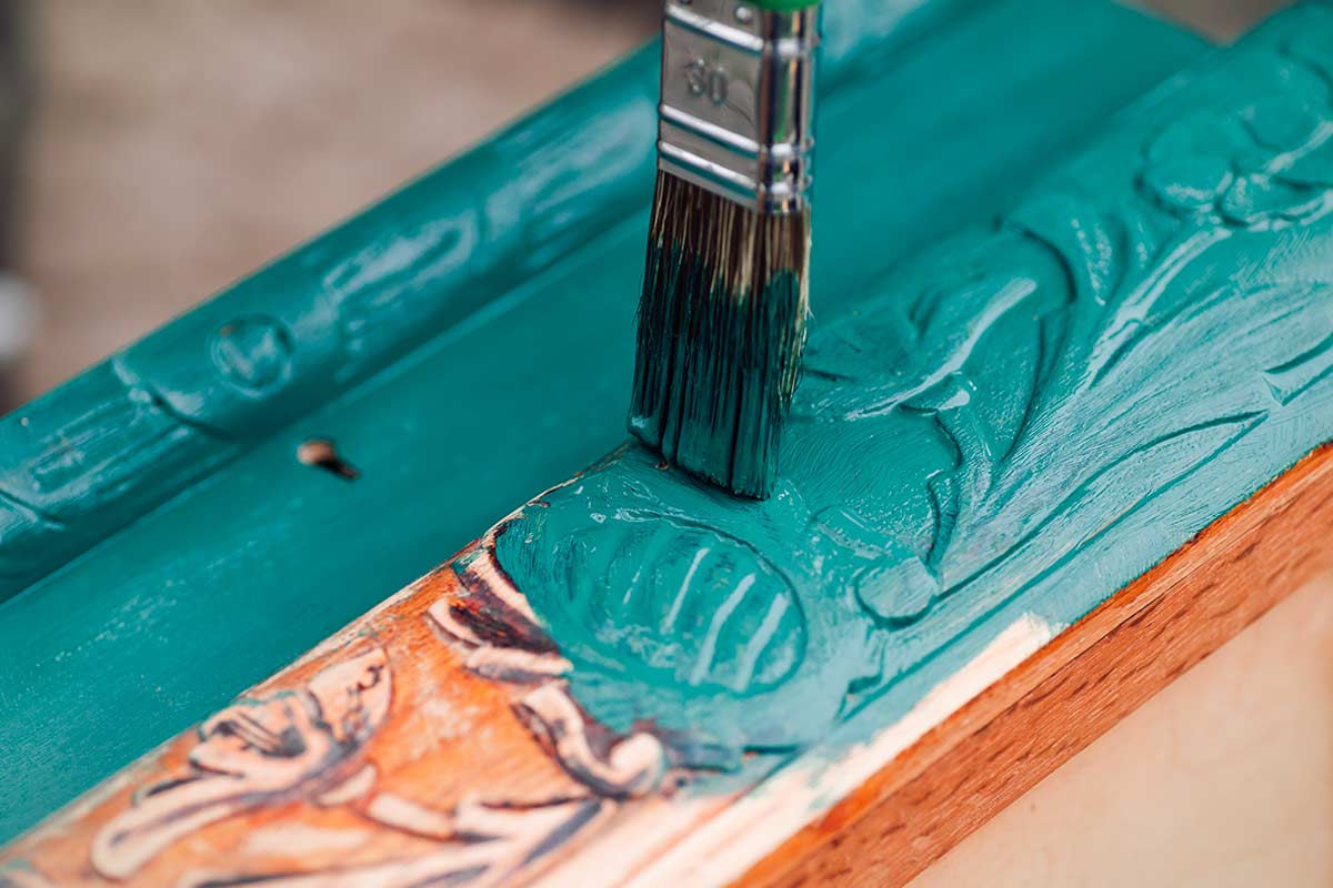 Pennello che stende vernice turchese su un mobile di legno intagliato, dettaglio del restauro fai da te.