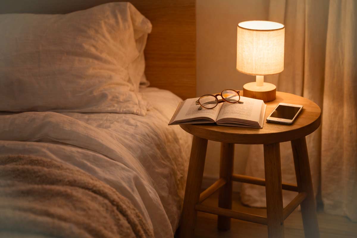 Sgabello in legno usato come comodino accanto al letto, con lampada accesa, libro, occhiali e telefono, in una camera dalla luce soffusa.
